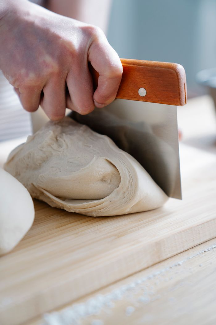 A hand slicing fresh bread dough with a metal cutter on a wooden board.